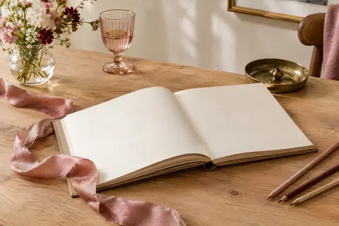 Livre d’or carré à couverture kraft ouvert sur ses pages vierges, posé avec un ruban vieux rose et des crayons