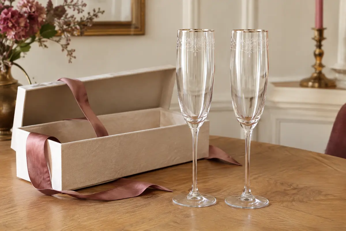 Deux flûtes à champagne en cristal gravé posées côte à côte devant leur coffret cadeau ouvert, sur une table en bois clair.