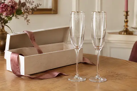 Deux flûtes à champagne en cristal gravé posées côte à côte devant leur coffret cadeau ouvert, sur une table en bois clair.