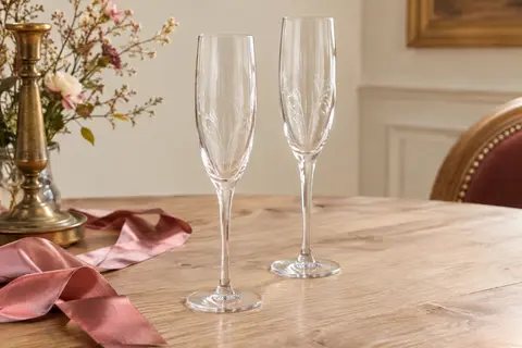 Deux flûtes à champagne en cristal gravé posées sur une table en bois clair avec un ruban vieux rose.